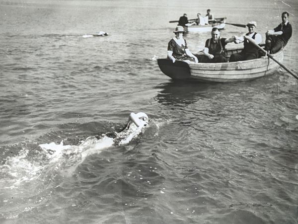 Black and white photo of two cross bay swimmers in the water, each following closely behind a rowing boat.